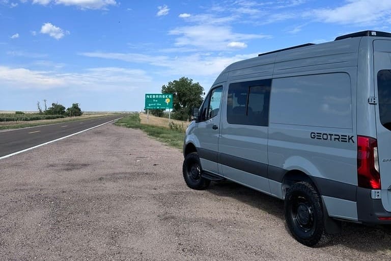 A sprinter camper van sits roadside next to a welcome to Nebraska sign.