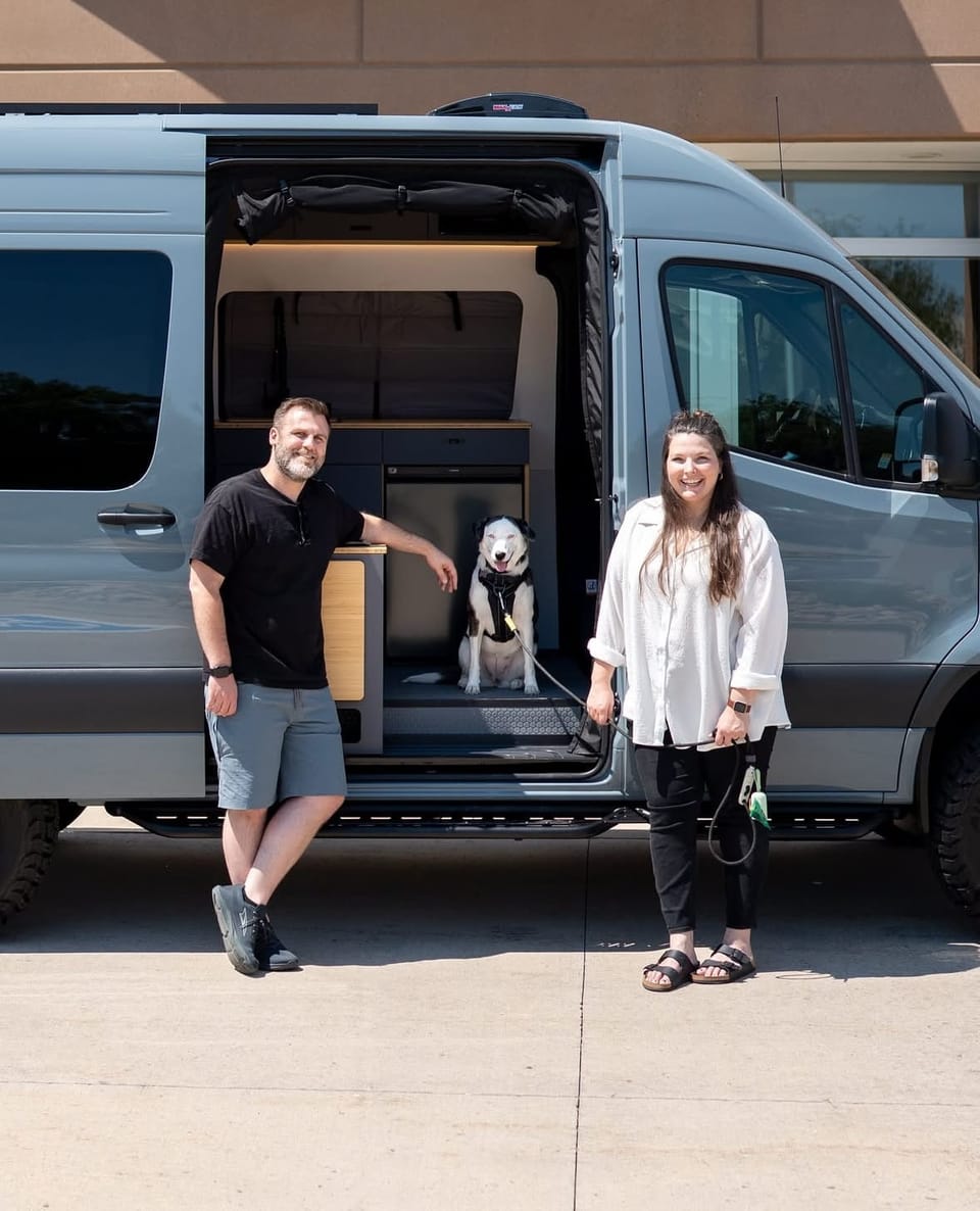 A man and a woman stand outside of a adventure campervan, the sliding door is open. A dog sits inside.